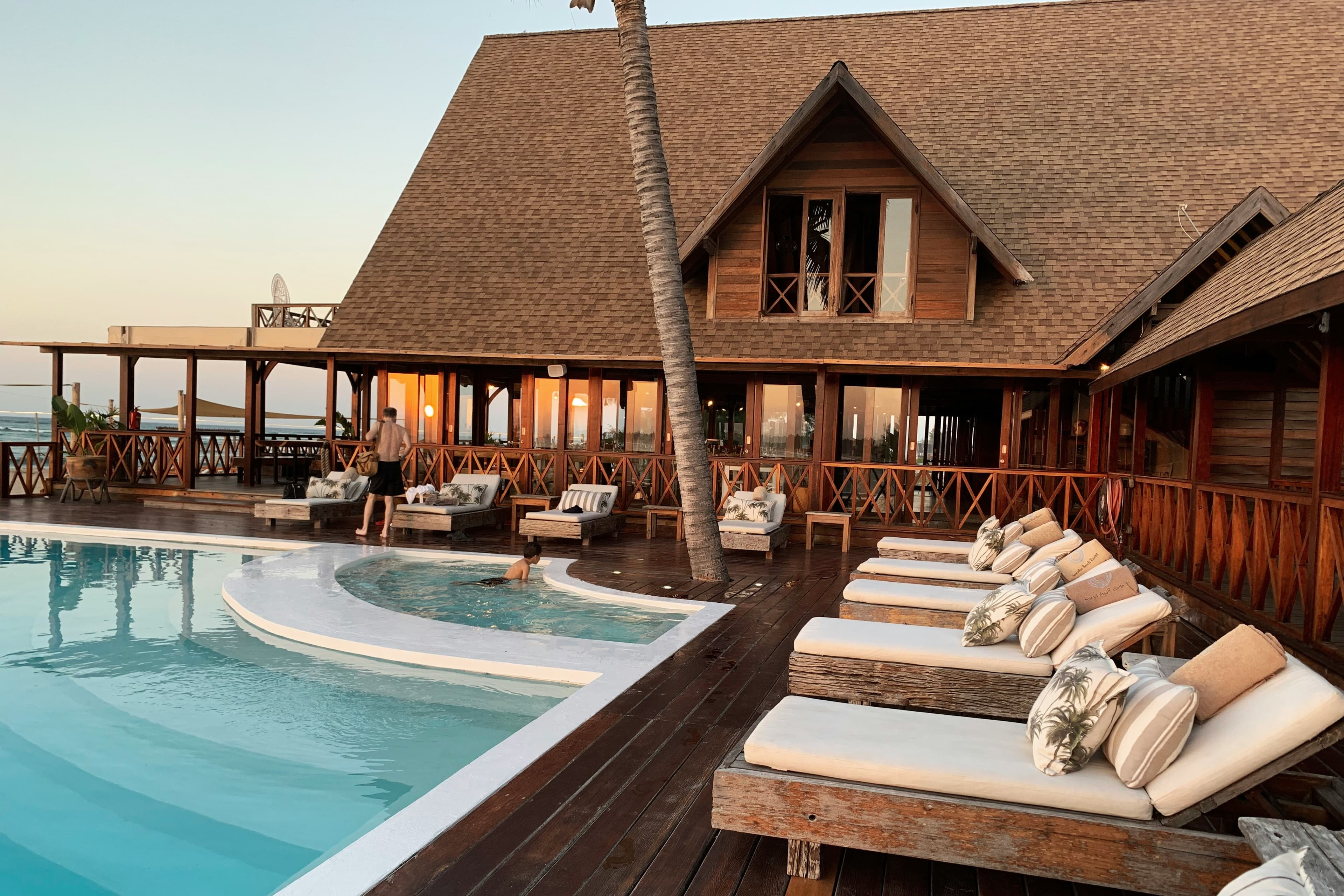 A lodge-style hotel with a thatched roof, terrace loungers and a pool at dusk.