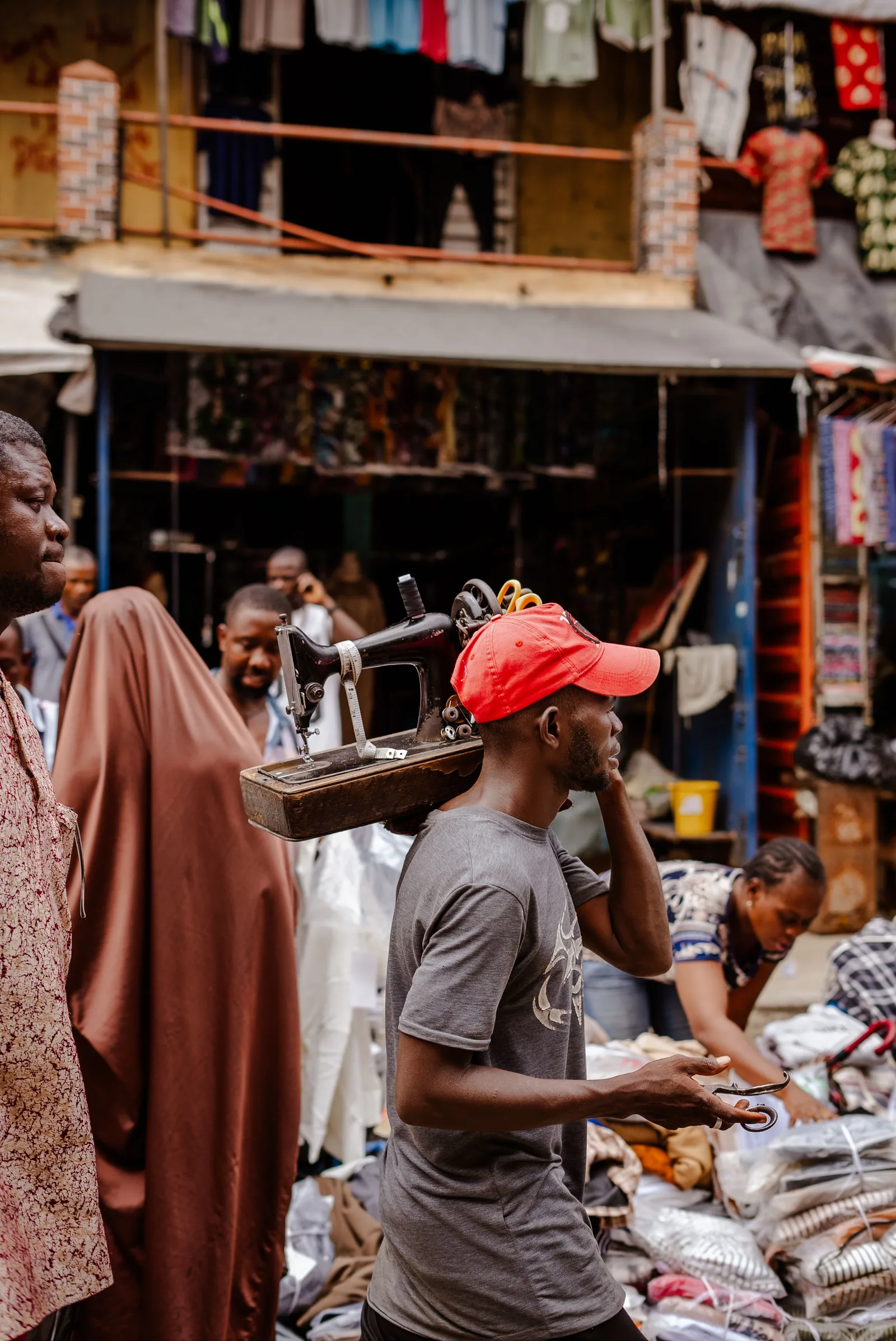 A tailor carrying a sewing machine through a busy African market street.