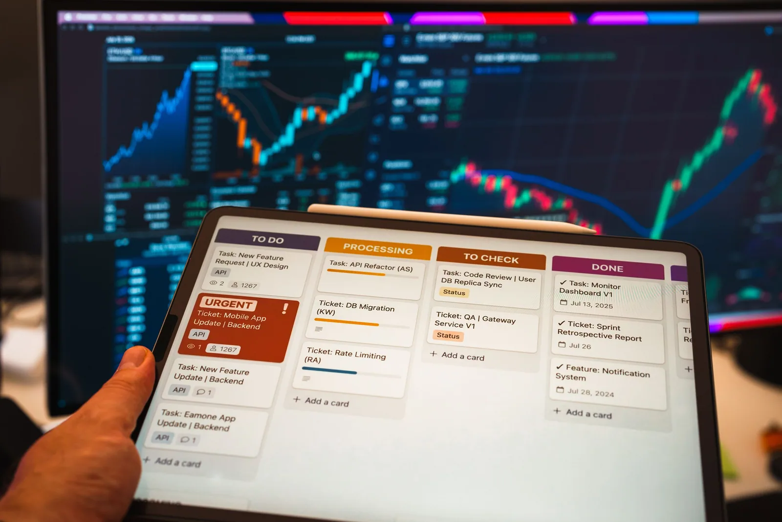 A hand holding a tablet showing a project task board, with market charts visible on a screen behind it.
